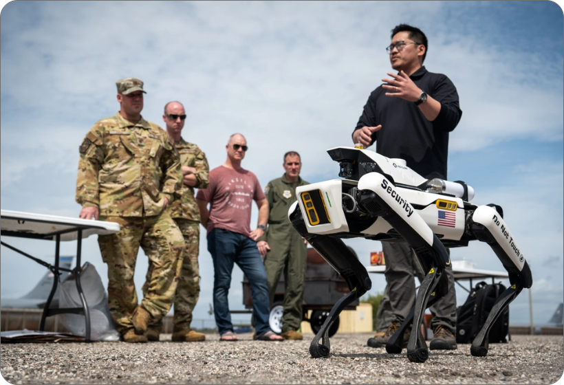 A man in a black shirt speaks while gesturing toward a white, four-legged robot security dog labeled "Security" and "Do Not Touch" with an American flag decal. Four military personnel, two in camouflage and two in civilian clothes, stand to the left, observing the presentation outdoors.