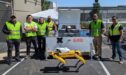 Six workers wearing high-visibility vests and sunglasses stand behind a yellow, four-legged security dronedog and a white mobile docking station labeled "GXO." A drone is visible resting on top of the docking station.