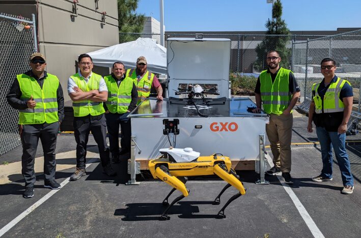 Six workers wearing high-visibility vests and sunglasses stand behind a yellow, four-legged security dronedog and a white mobile docking station labeled "GXO." A drone is visible resting on top of the docking station.