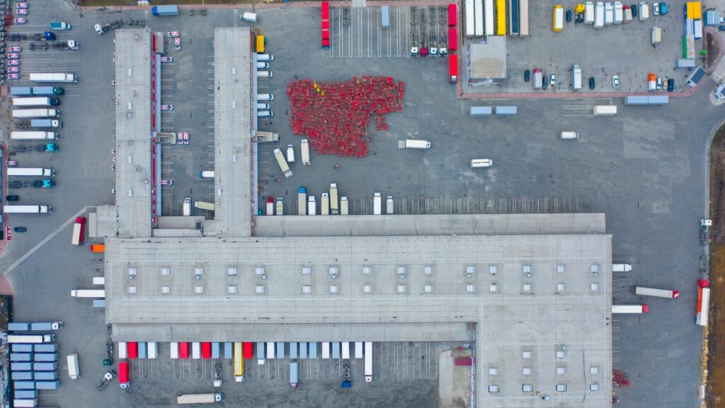 An aerial top-down view shows a large logistics park with a warehouse and semi trucks at loading docks.