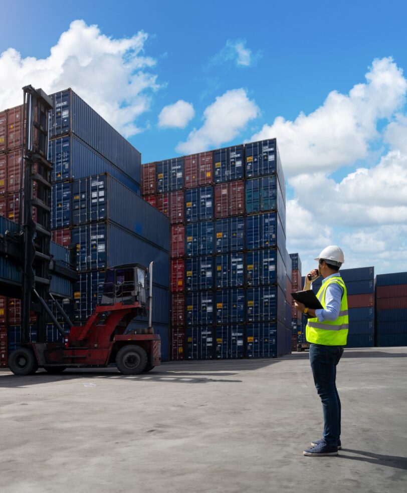 A man in a white hard hat and a high-visibility vest stands in a shipping yard, speaking into a handheld radio while holding a clipboard.