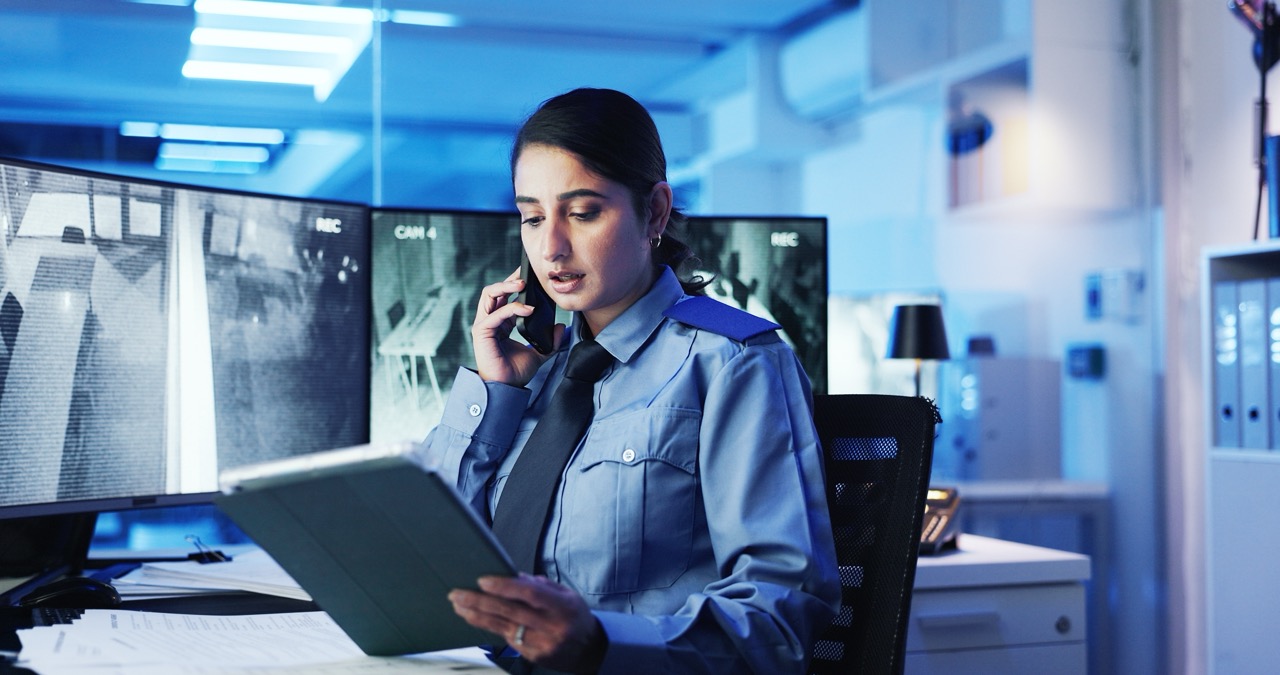 Security officer sitting at cubicle with CCTV monitors holds tablet computer and talks on her phone, fighting off security guard fatigue. 