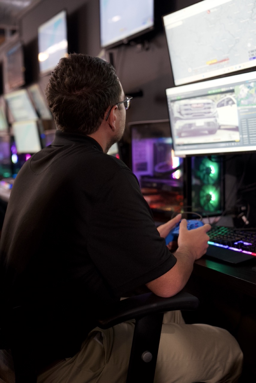 Skilled robotics operator sits at monitor in remote security operations center, reviewing real-time video feed from ground robot. 
