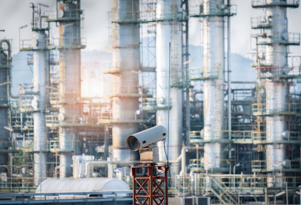 Fixed surveillance camera at an oil refinery, several tall flare stacks and scaffolding in background.