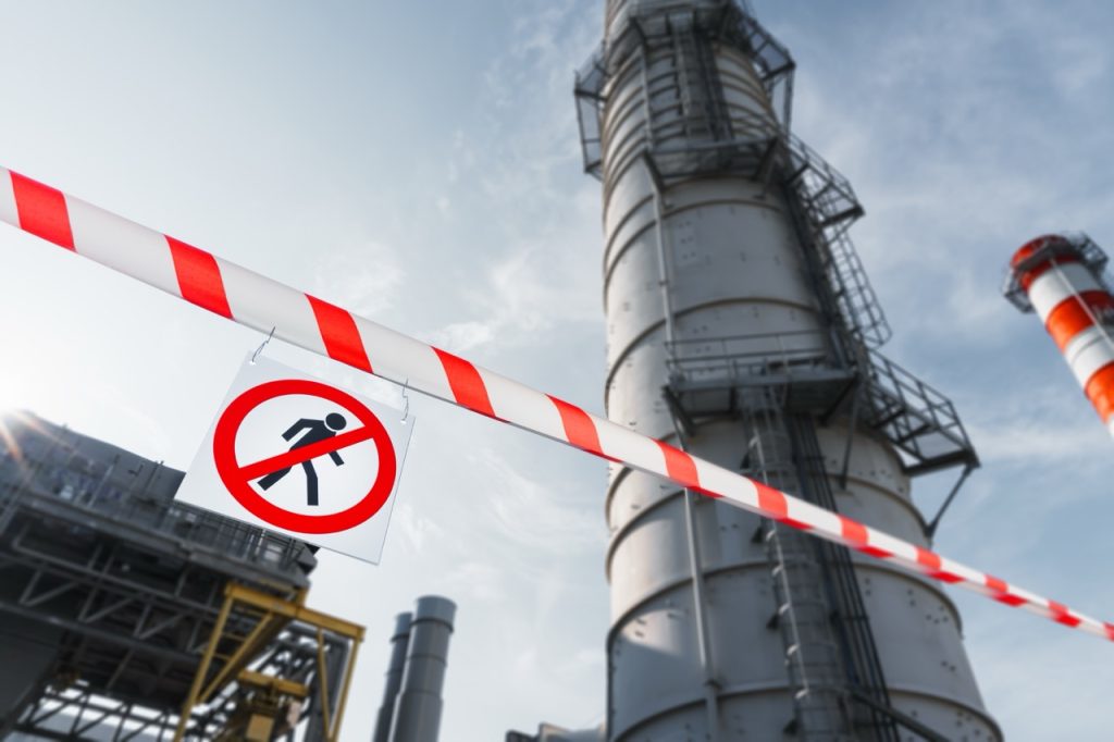 Sign indicating restricted access hangs from security tape at an oil refinery, flare stacks in background.
