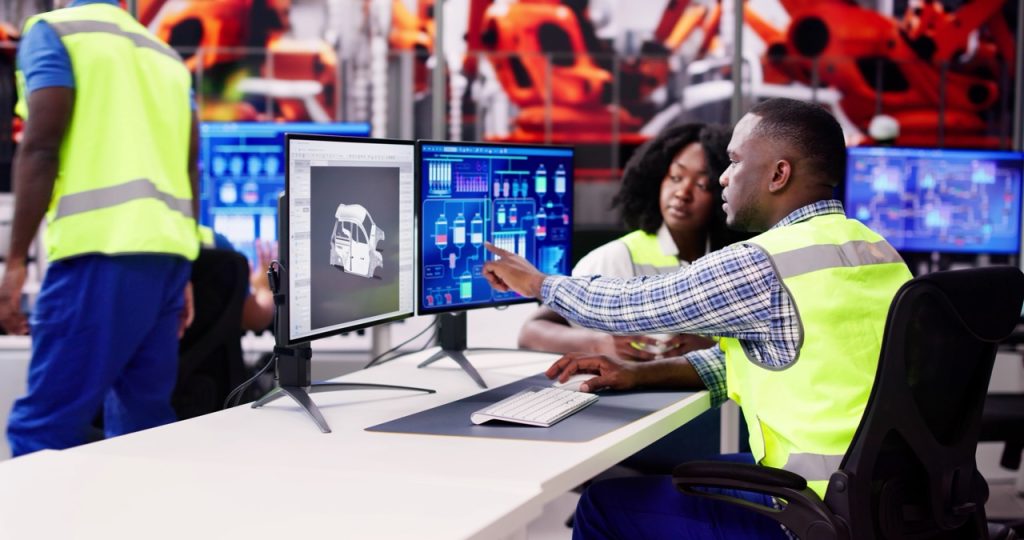 Security operator at chemical plant sits in front of double computer monitor, discussing potential alert response with colleague.