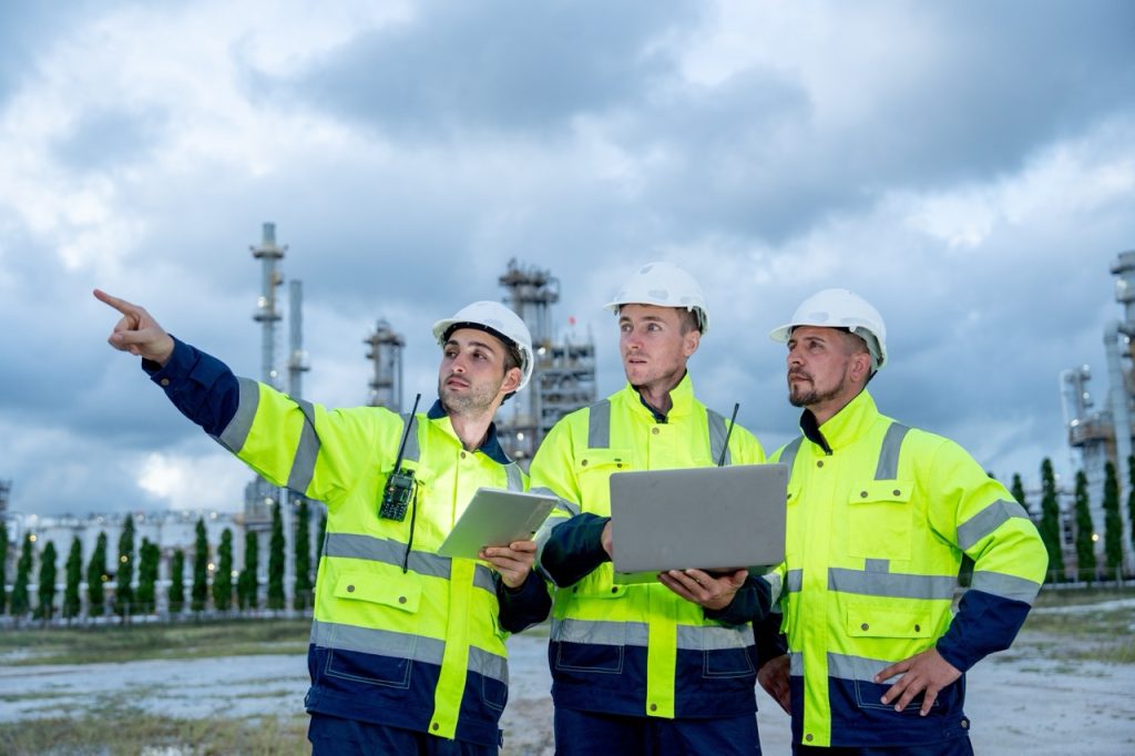 Three security inspectors in safety jackets and helmets review an incident at a chemical plant, holding laptop computers.