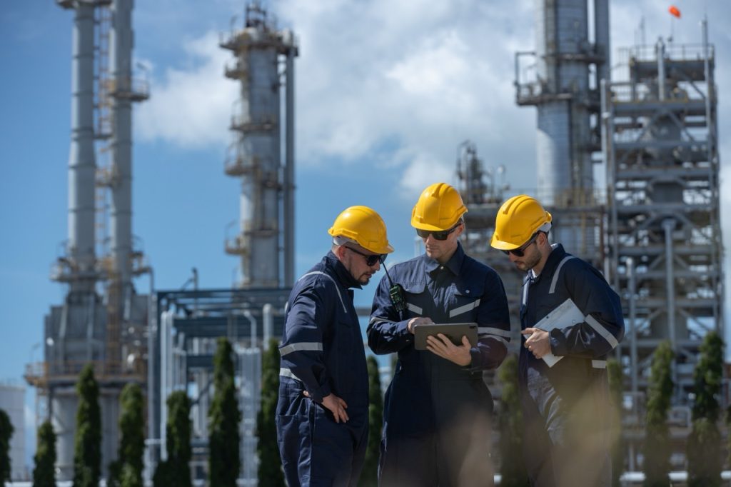 Three security engineers in hard hats consult using a tablet computer at an oil refinery, flare stacks and piping in background.