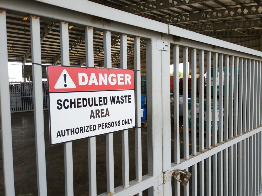 Metal fence secured with padlock, sign on fence reads, “DANGER - SCHEDULED WASTE AREA - AUTHORIZED PERSONS ONLY”