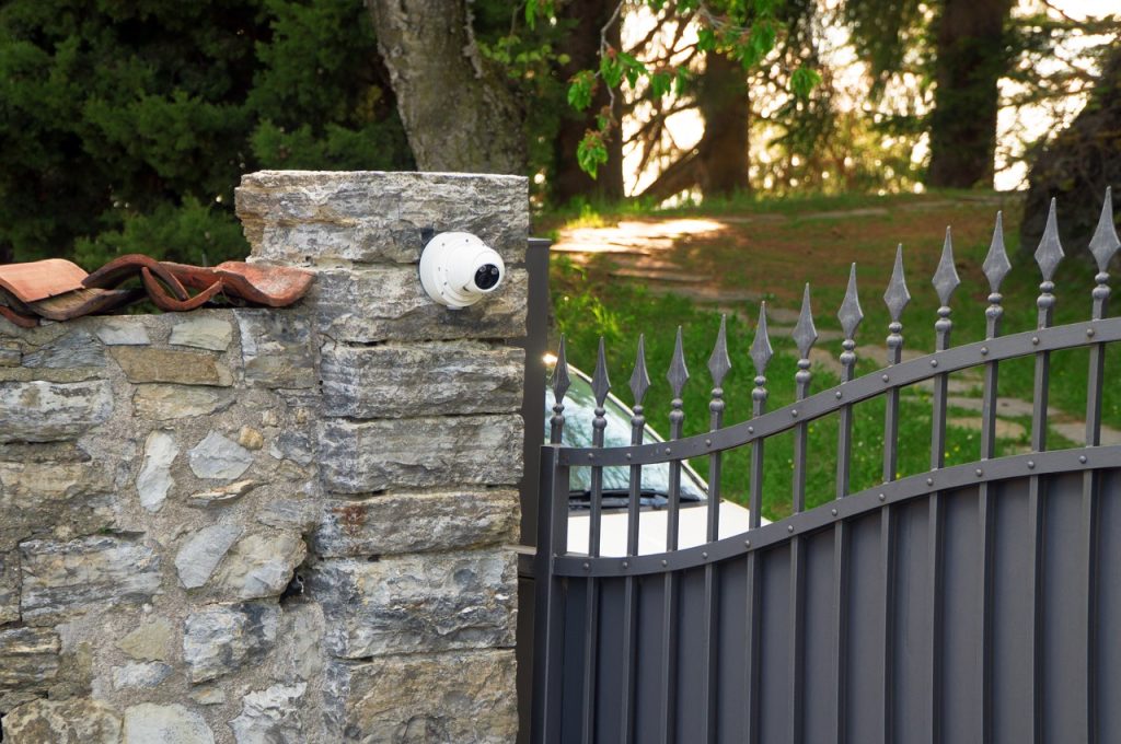 Private estate entrance with a gated driveway, stone gateposts, and perimeter security camera monitoring property access.