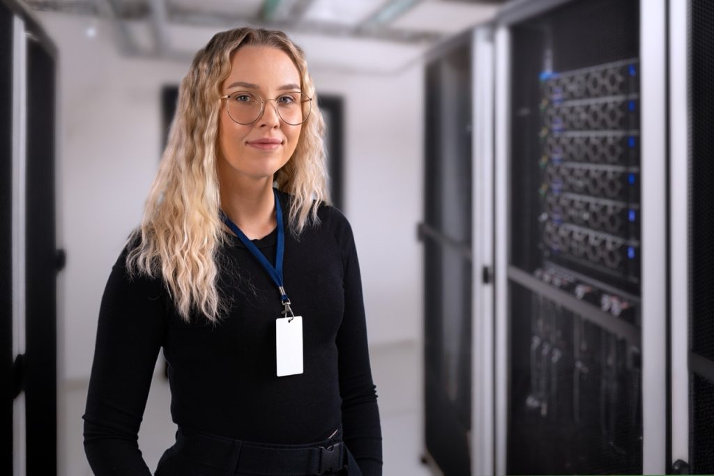 Data center professional wearing access badge on lanyard stands beside server racks in secure server room.