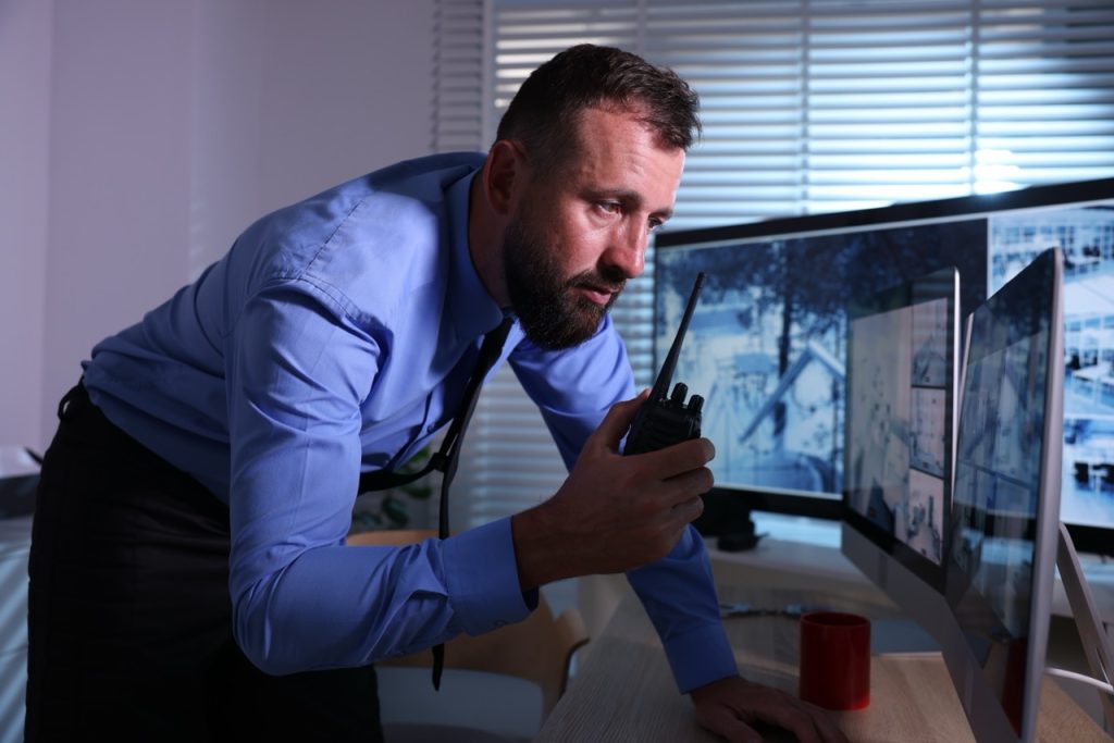 Security operator uses two-way radio while monitoring construction site surveillance camera feeds on multiple monitors.