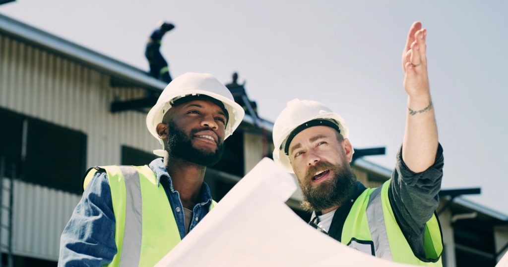 Two construction site managers in hard hats and safety vests review plans and survey a jobsite.
