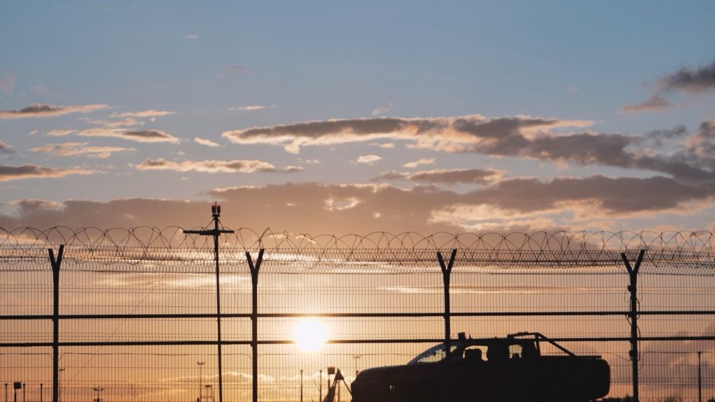 Airport perimeter fence and security vehicle monitoring the airfield at sunset. 