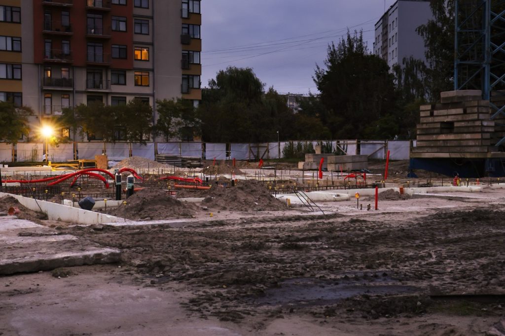 Construction site at dusk with exposed foundations, piping, stacked materials, temporary fencing, and nearby apartments.  