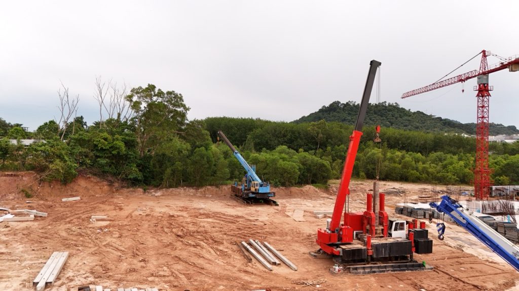 Construction site with mobile cranes, a tower crane, building materials, and cleared ground surrounded by trees. 