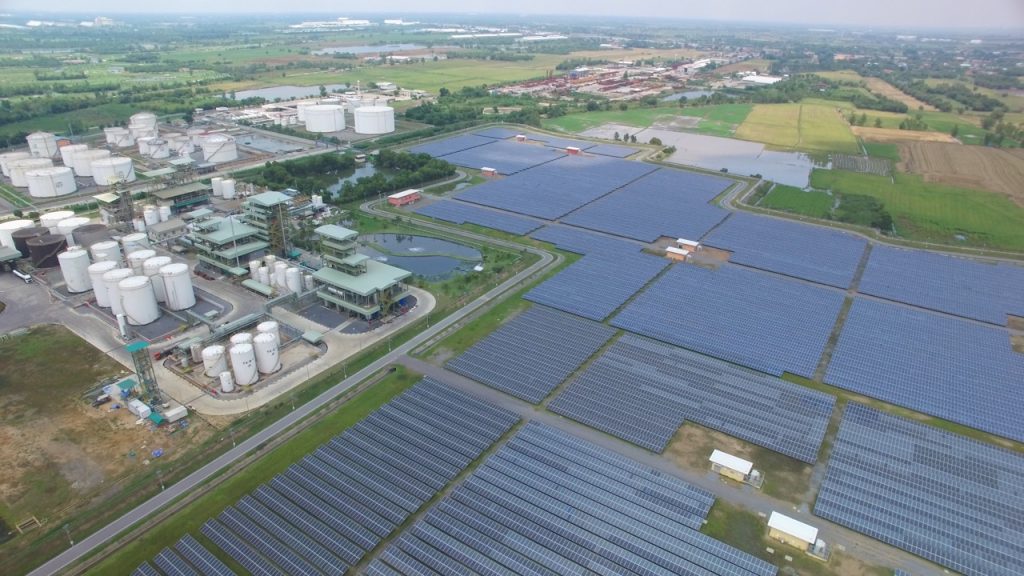 Aerial view of a solar farm beside an industrial energy facility with expansive grounds and perimeter roads. 