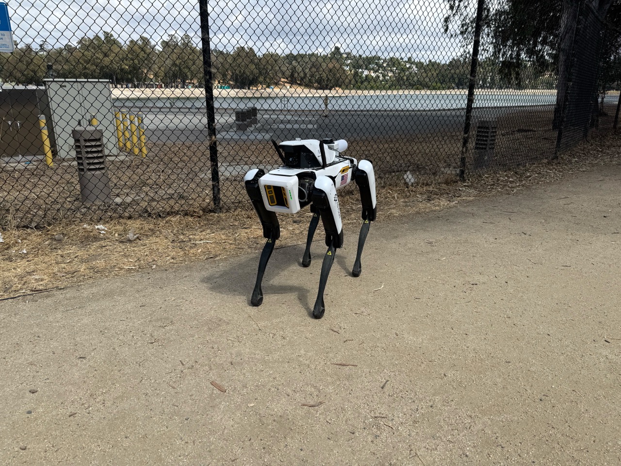 DroneDog patrolling a water treatment facility on a paved path next to a chain-link fence, with a body of water and facility infrastructure visible in the background. 