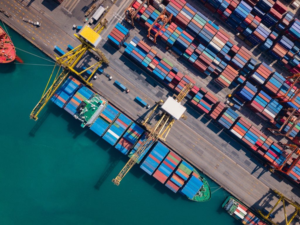 Aerial view of a maritime container terminal with stacked cargo, cranes, loading berths, and adjacent waterfront.