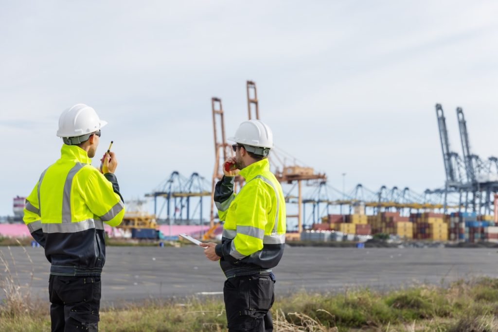 Two maritime terminal security personnel in high-visibility jackets communicate by radio near container cranes and cargo stacks.