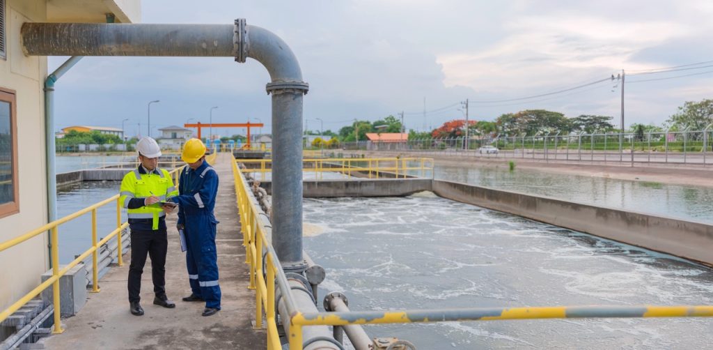 Two workers inspect equipment beside settling basins at a water treatment plant with pipelines, walkways, and perimeter fencing in the background. 