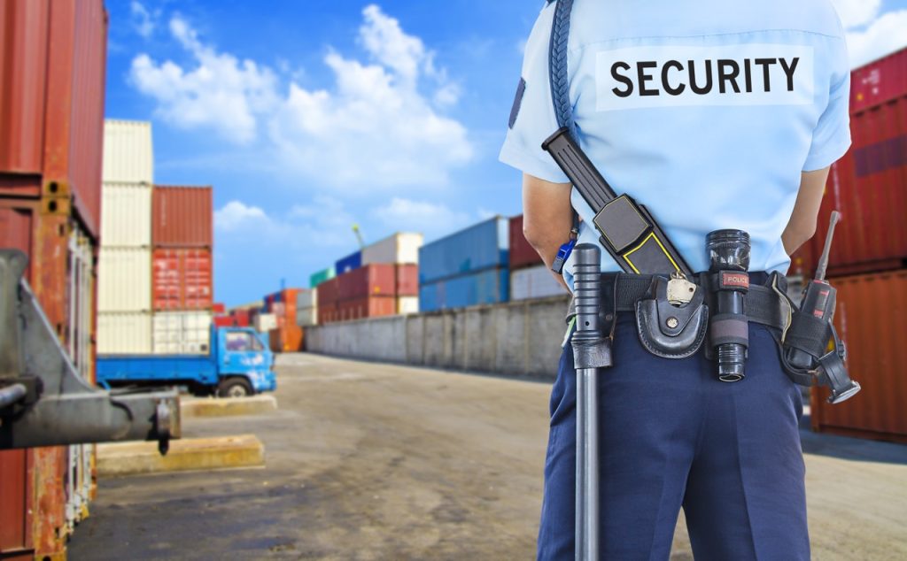 Heavily armed security officer stands with back to camera, watching stacked cargo containers at a maritime terminal yard.