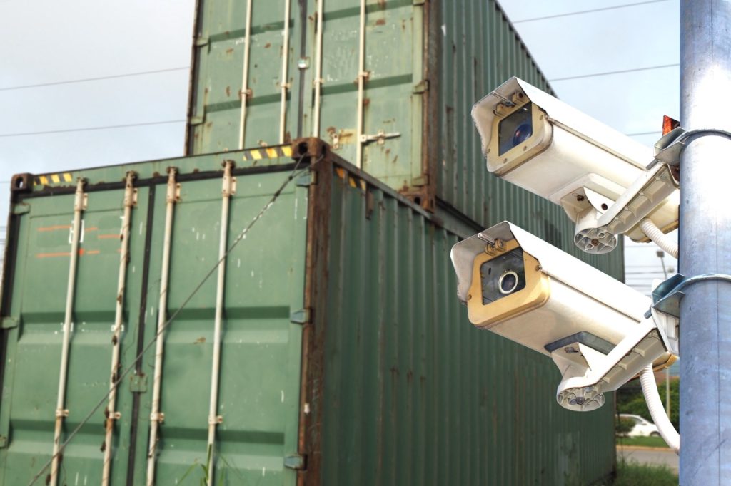 Two fixed security cameras monitor stacked cargo containers at a maritime terminal yard.