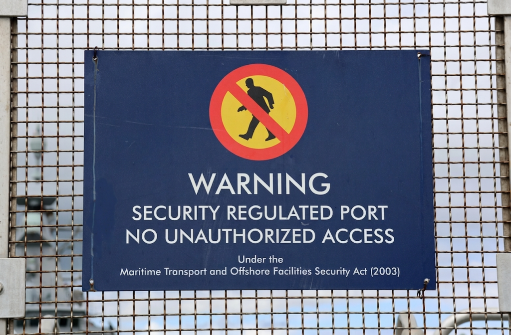 Sign affixed to metal fence at maritime terminal reads “WARNING. SECURITY REGULATED PORT. NO UNAUTHORIZED ACCESS”